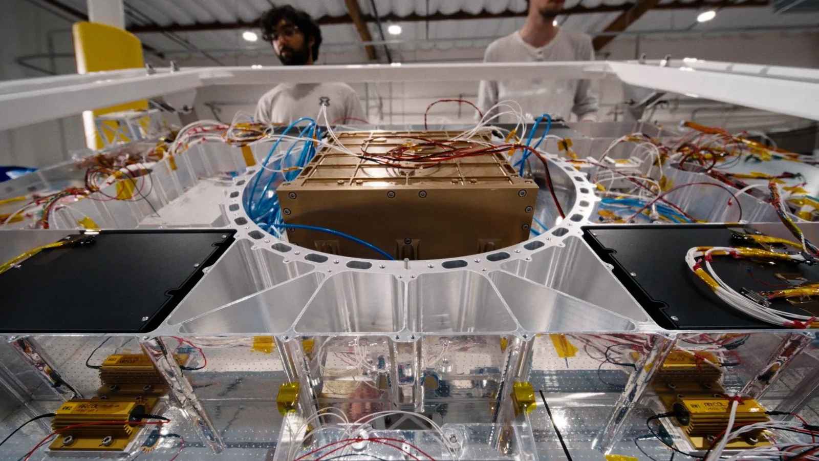 Two people in light-colored shirts look over a large aluminum spacecraft-style structure in a workshop. A gold-colored rectangular component sits in its center.